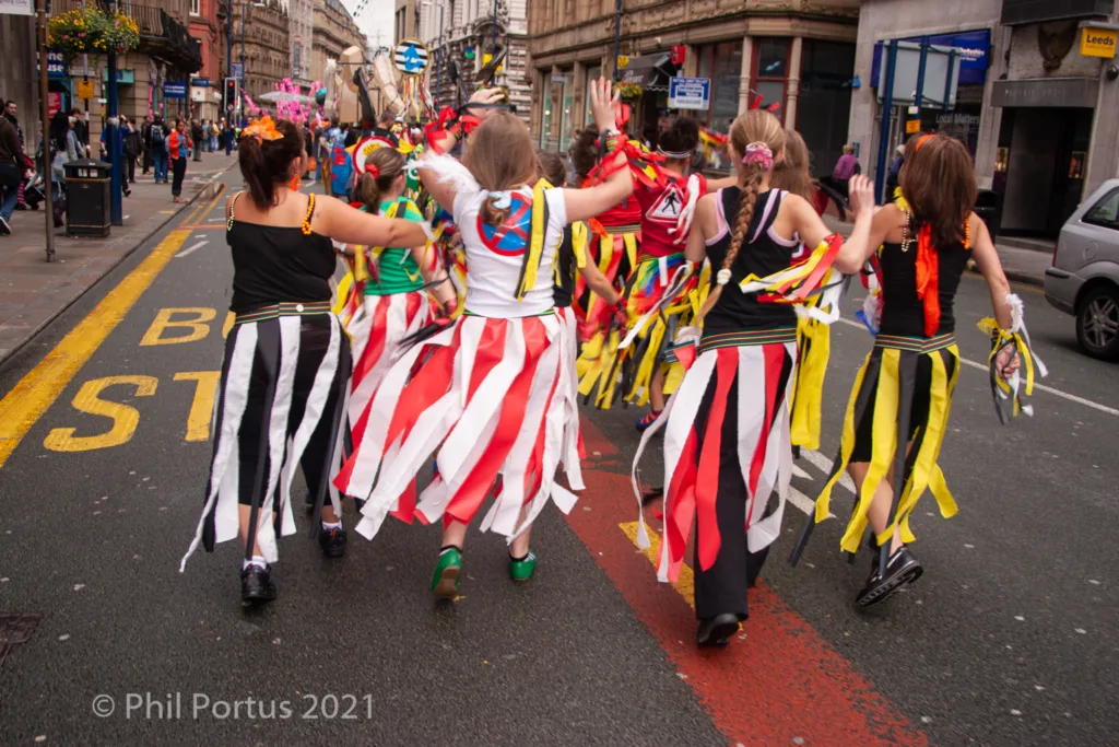 A group of people wearing colorful costumes with ribbons dance down a street lined with buildings and pedestrians.