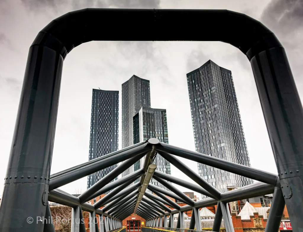 A metal arch frames a view of three tall, dark buildings against an overcast sky.