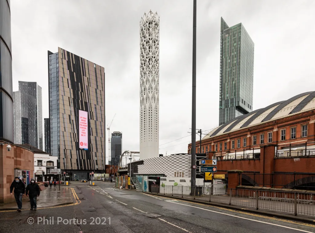 Tall modern buildings line a wet street with a lamppost and brick structures visible in the background.