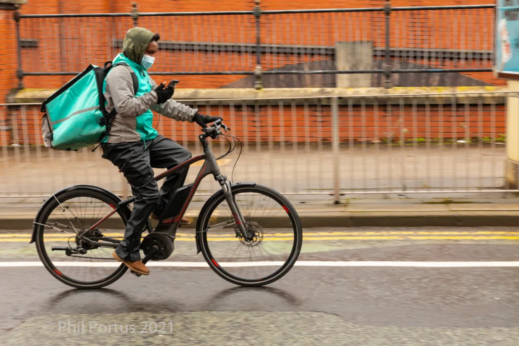 A person on an electric bicycle rides down a wet street with a large delivery bag and face covering.