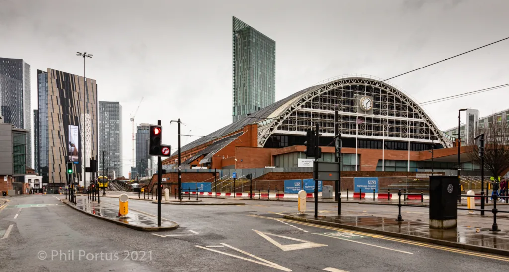 A wide street leads toward a large arched building flanked by modern high-rise structures under an overcast sky.