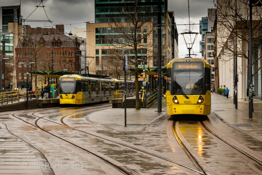 Two yellow trams curve along wet tracks in a city setting with modern buildings and bare trees.