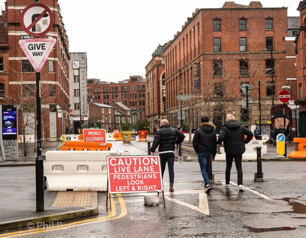 Three people walk across a street with construction barriers and signage in an urban environment.