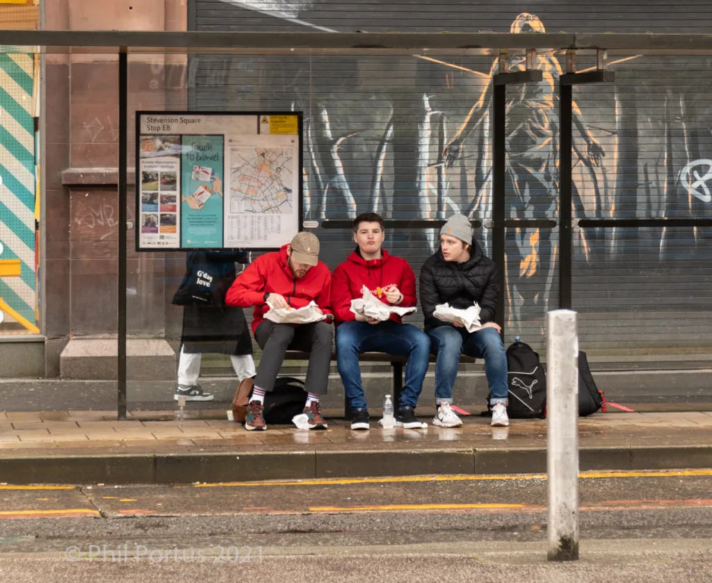 Three people sit on a bus stop bench eating food while waiting outdoors.