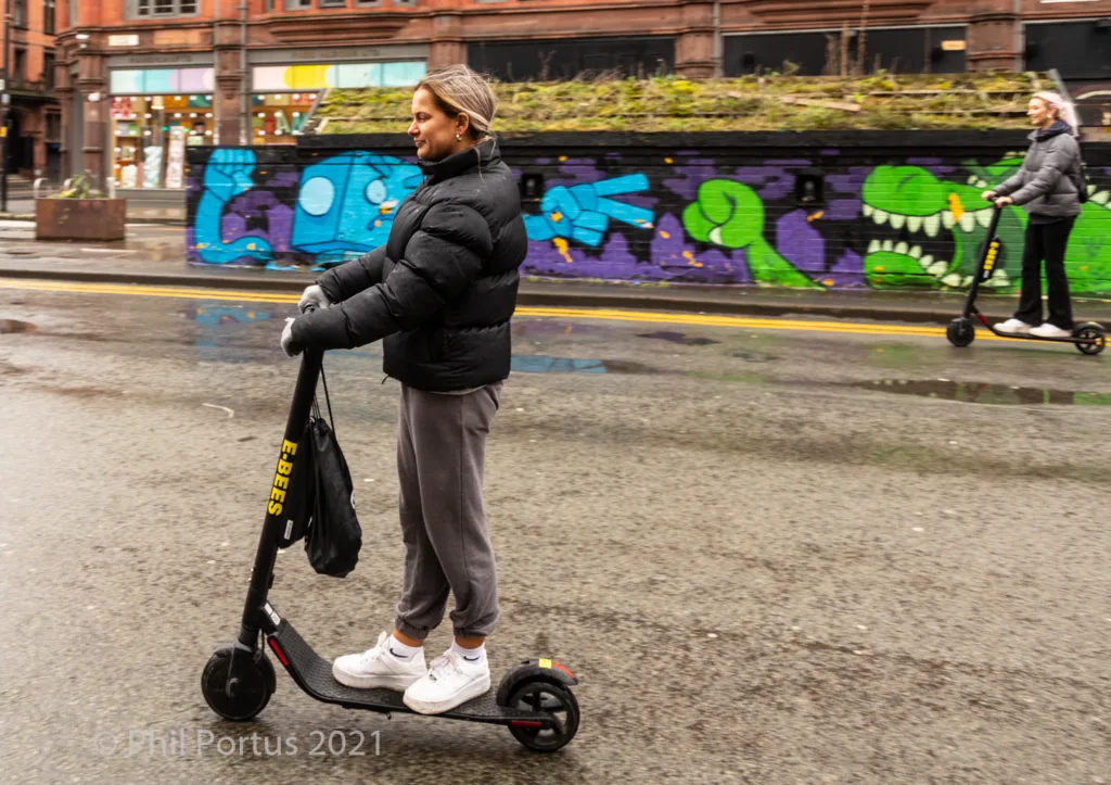 A person on an electric scooter stands on a wet street with graffiti and buildings in the background.