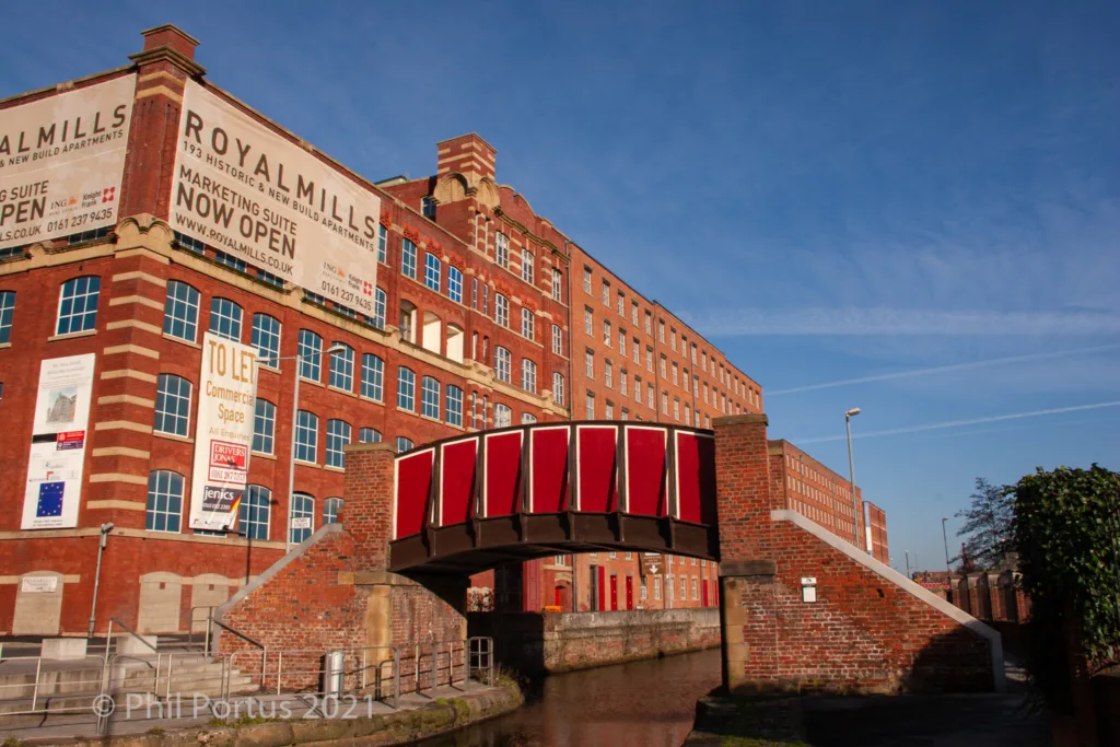 A red footbridge spans a waterway in front of a large brick building with multiple windows.