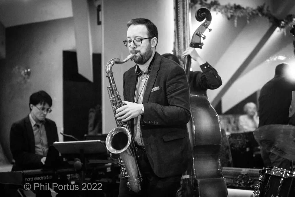 A musician plays a saxophone while other band members perform in black and white.