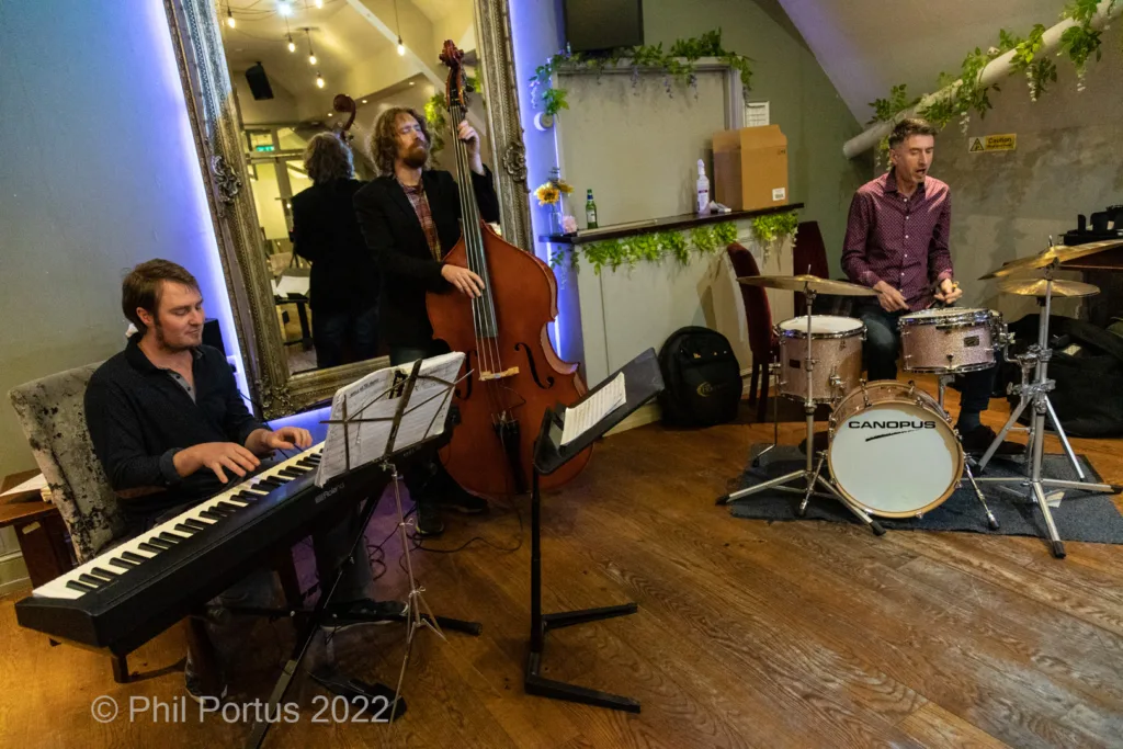Four musicians perform on a wooden stage with instruments including keyboard, bass, and drums.