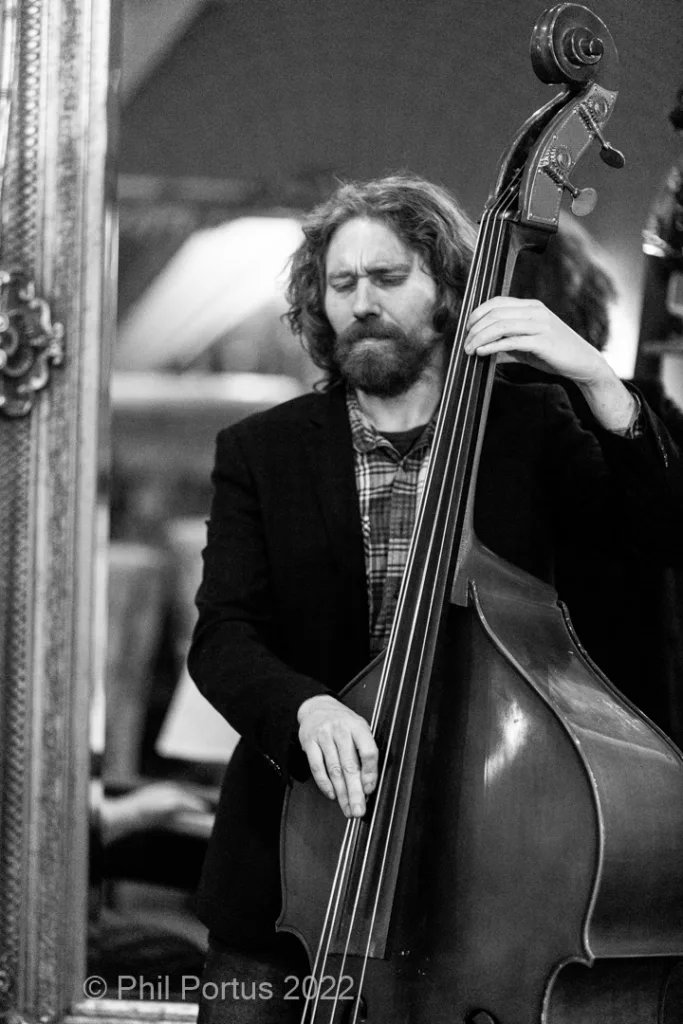 A musician with a beard plays an upright bass in black and white.