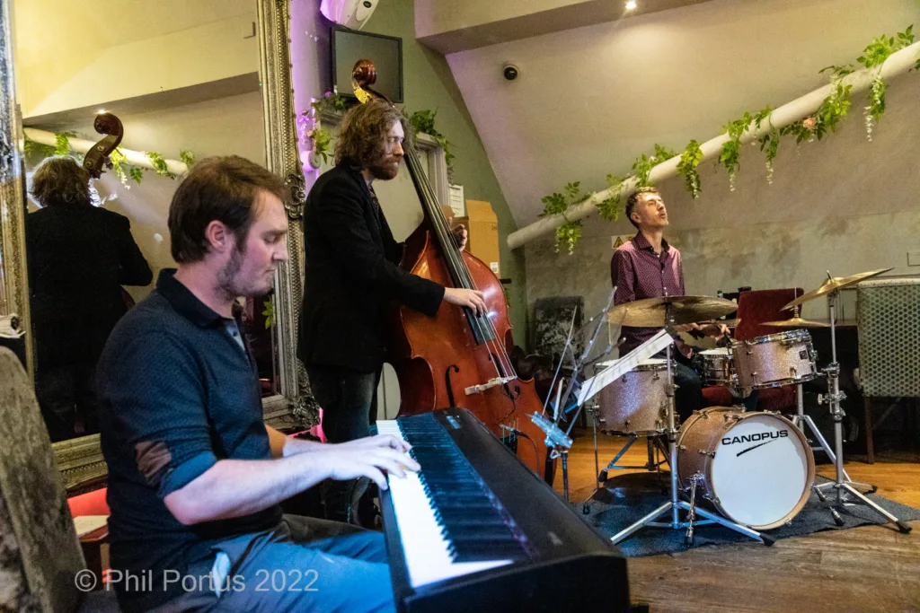Four musicians perform on stage with a piano, bass, and drum set in a dimly lit room.
