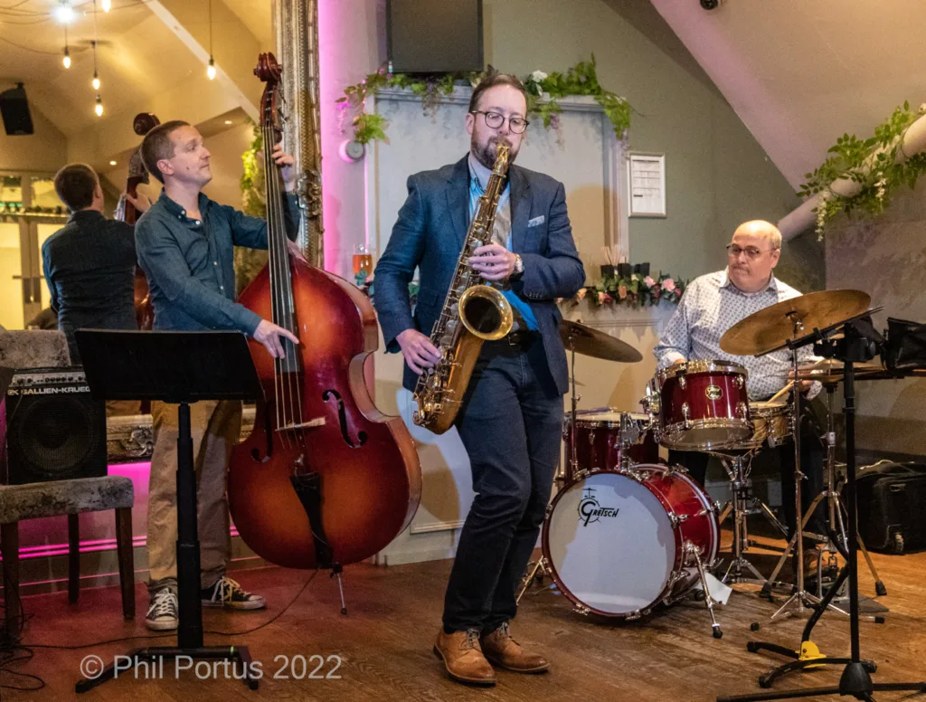 Four musicians perform on a stage with instruments including a saxophone, double bass, keyboard, and drum kit.