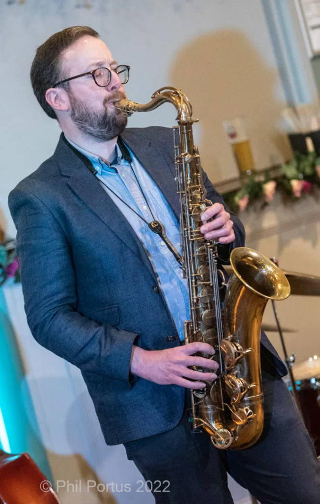 A musician wearing glasses plays a saxophone while dressed in a suit and collared shirt.