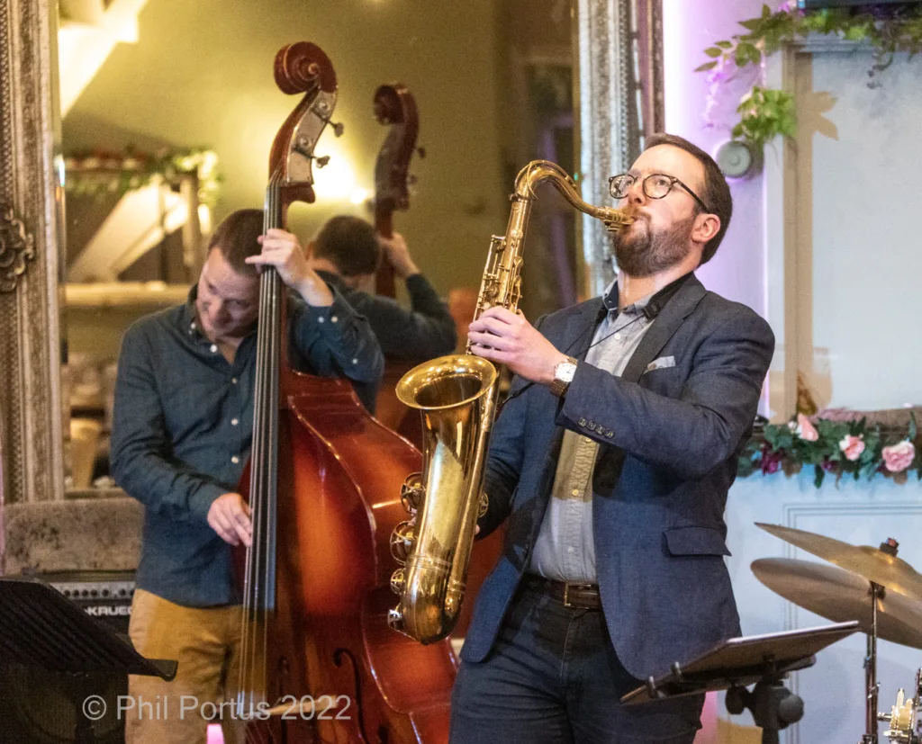 A musician plays a saxophone while another plays an upright bass in what appears to be a formal room.