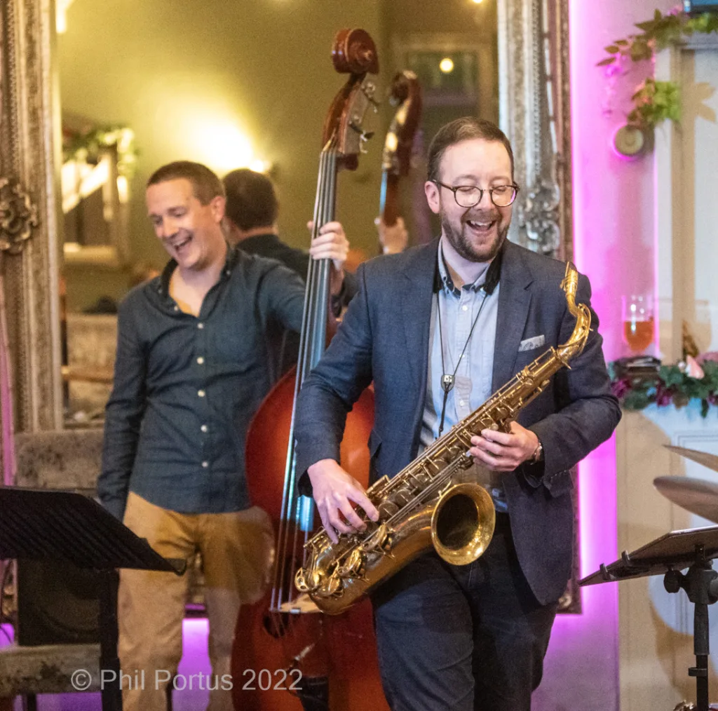 Three musicians perform on stage during a concert with pink lighting and decorative plants visible in the background.