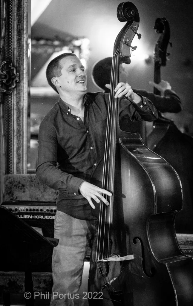 A musician plays a double bass while smiling, standing near a piano in black and white.