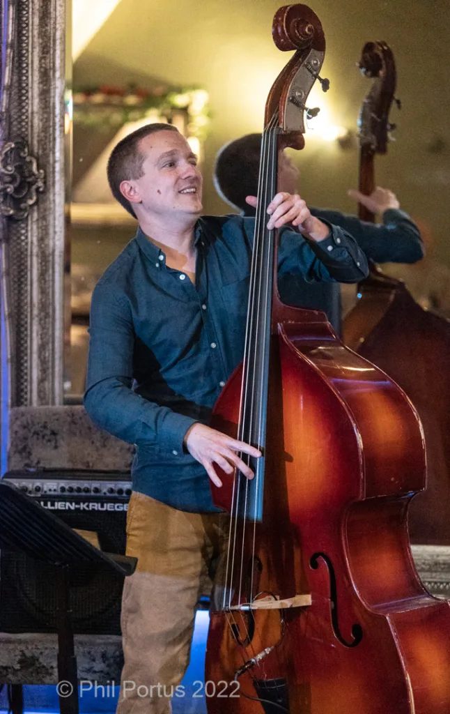 A musician plays a double bass on stage, smiling while interacting with another performer.