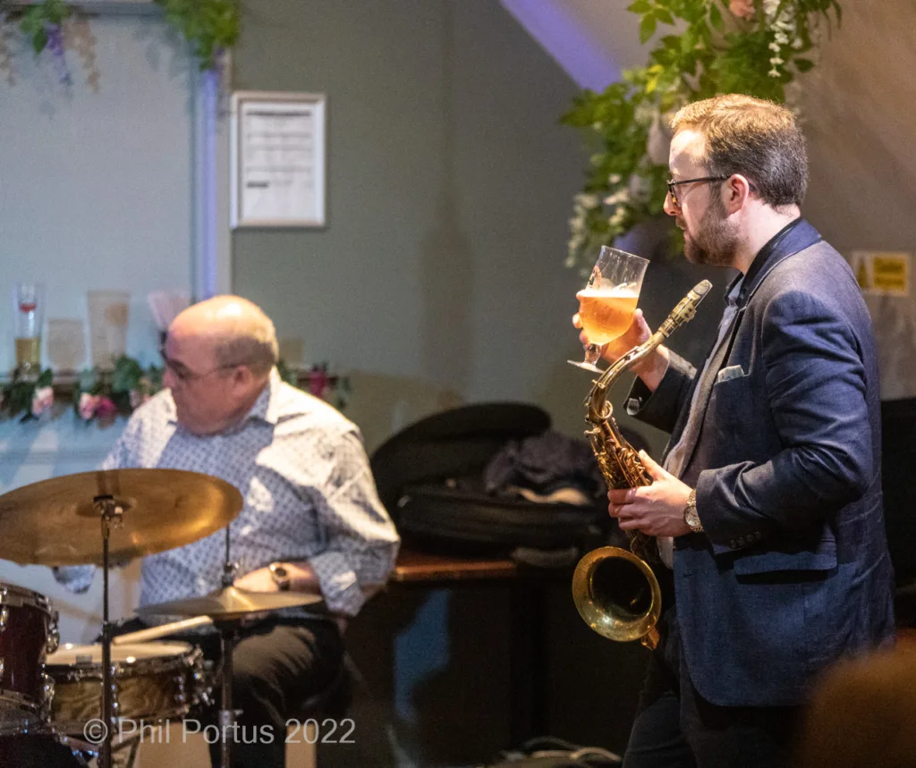 A musician plays a saxophone while holding a drink, with a drummer visible in the background.