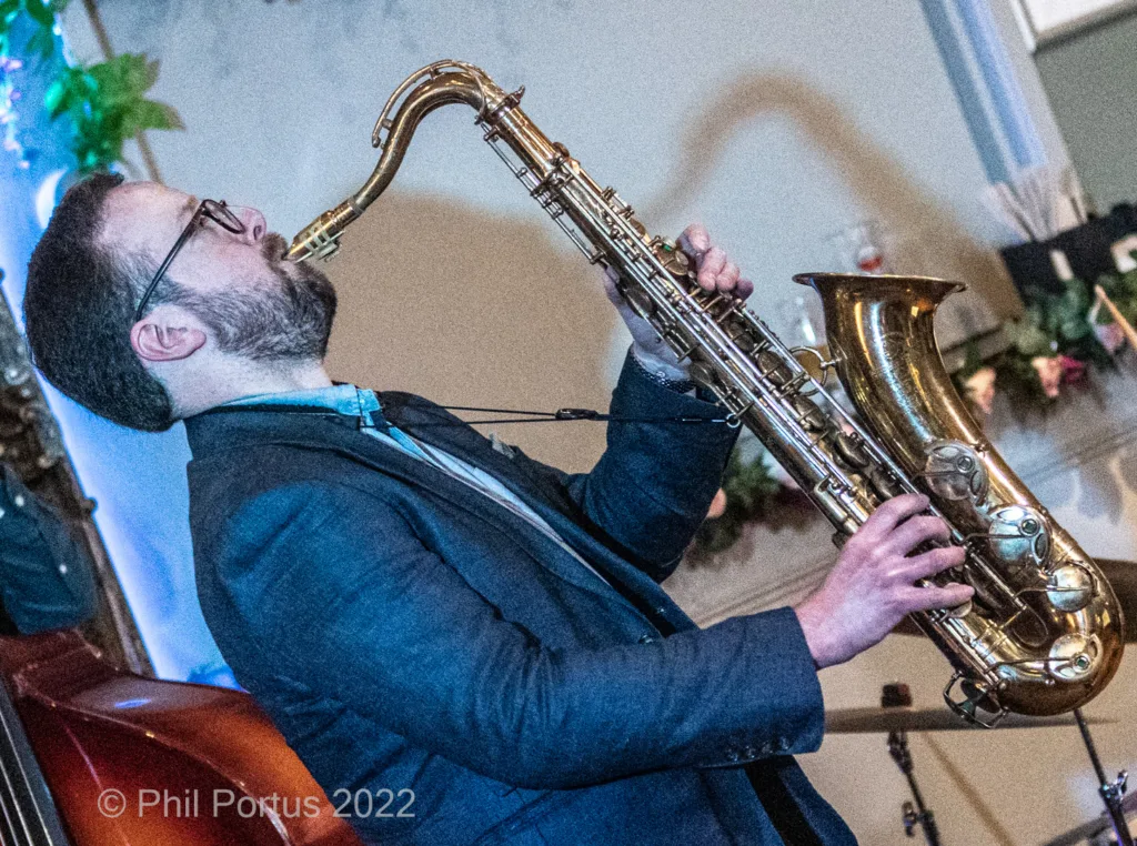 A musician with a beard plays a saxophone while wearing a dark suit and glasses.