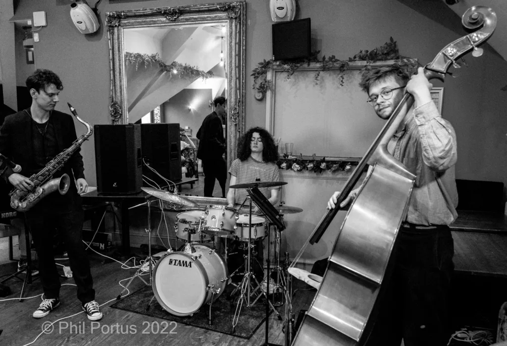 Three musicians perform on stage with a saxophone, drums, and upright bass in black and white.