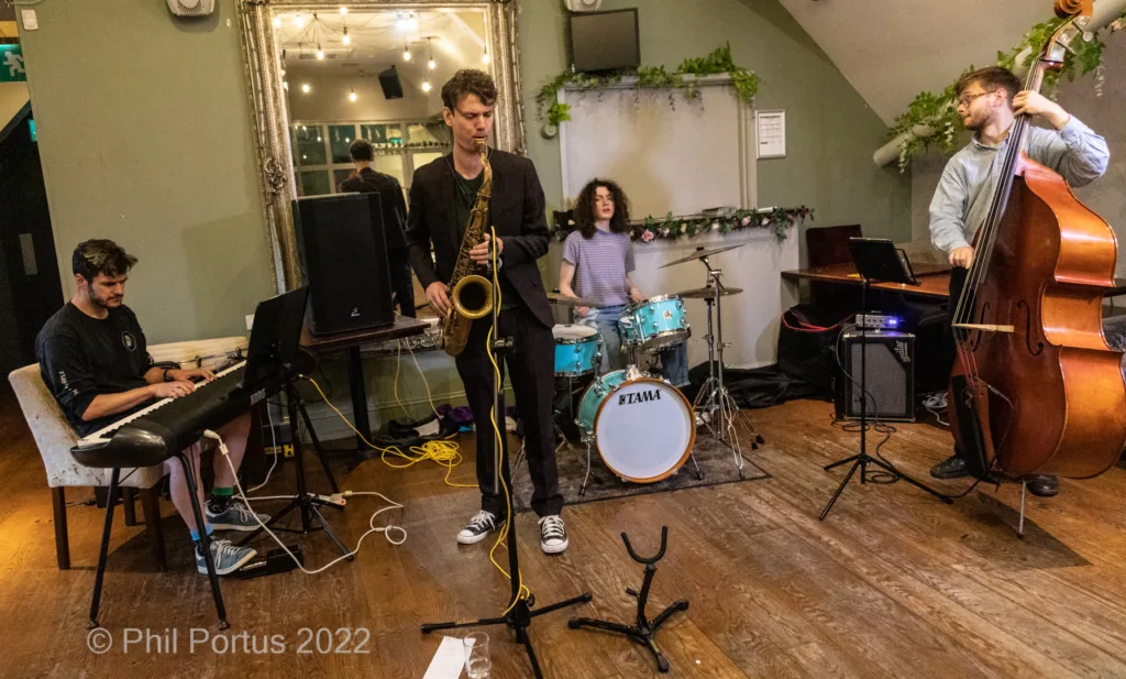 Four musicians perform on a wooden stage with instruments including keyboard, saxophone, drums, and upright bass.