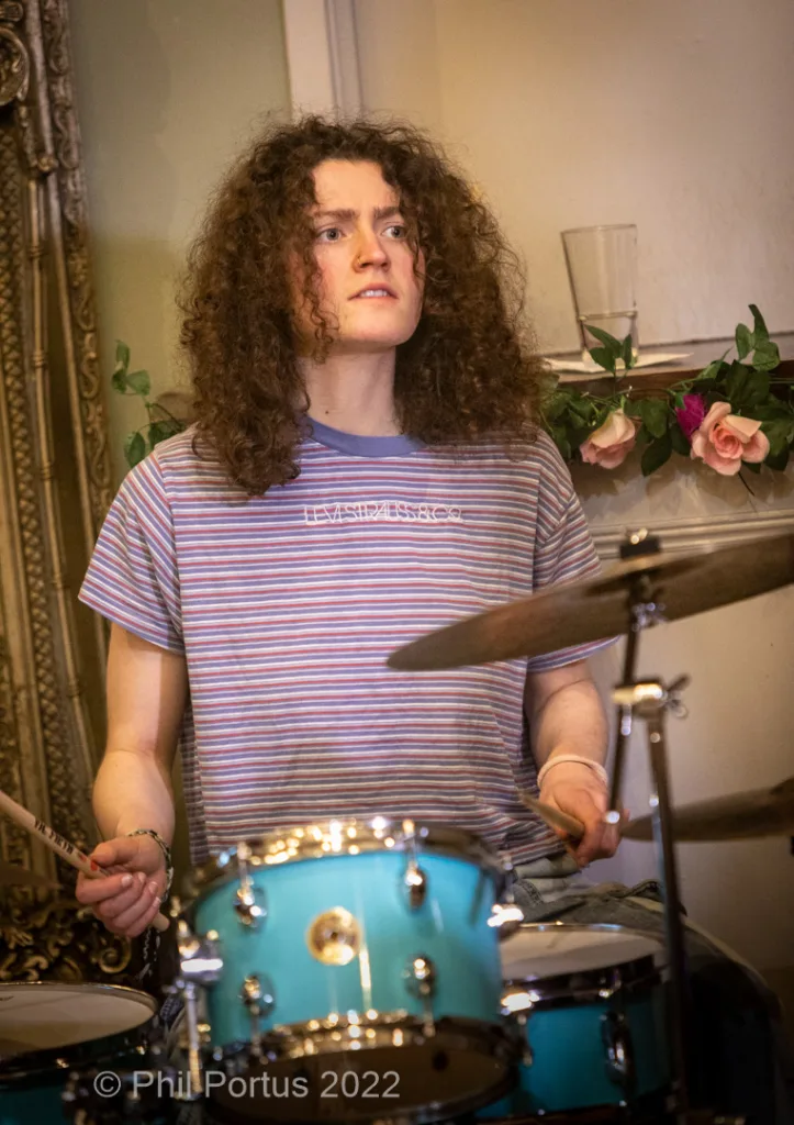 A person with curly hair plays a blue drum kit in a room with floral decorations and patterned curtains.