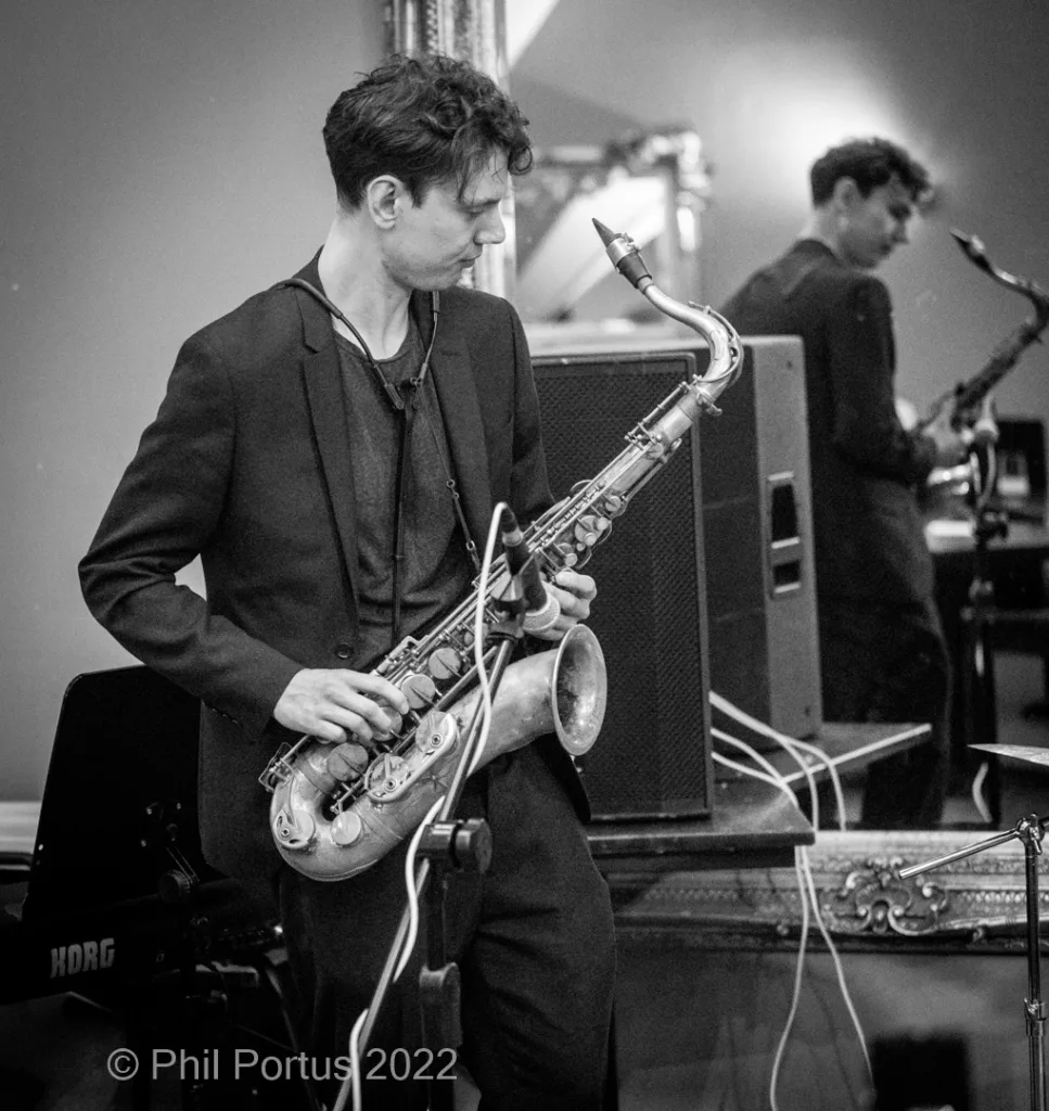 A musician in a dark suit plays a saxophone while another person is visible behind him at an event.