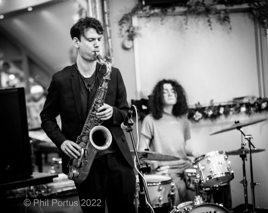 A musician plays a saxophone while another provides percussion on drums in black and white.