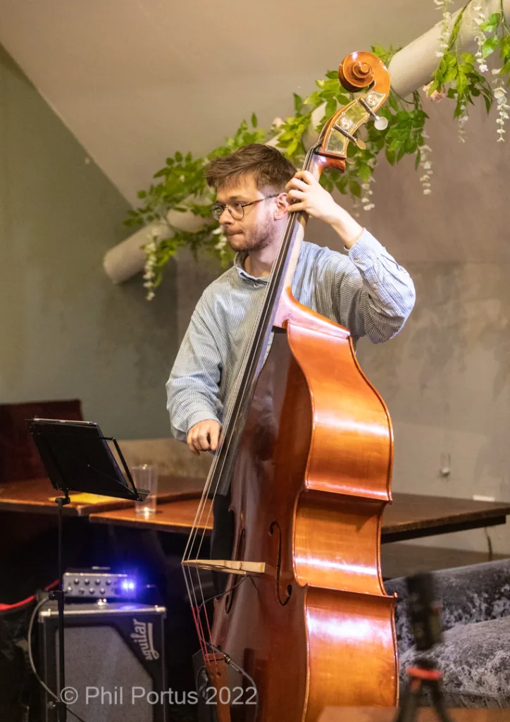 A musician plays an upright bass while wearing glasses and a blue shirt.