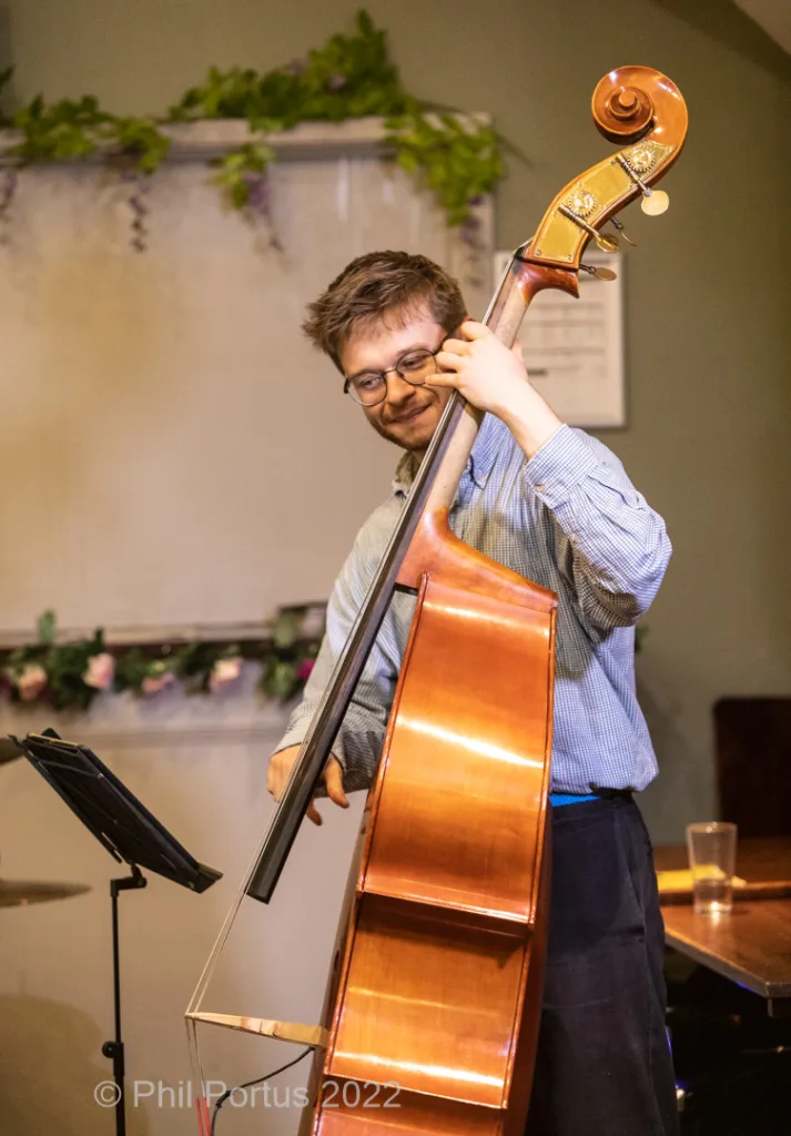 A musician with glasses plays a double bass in a room with greenery and tables.