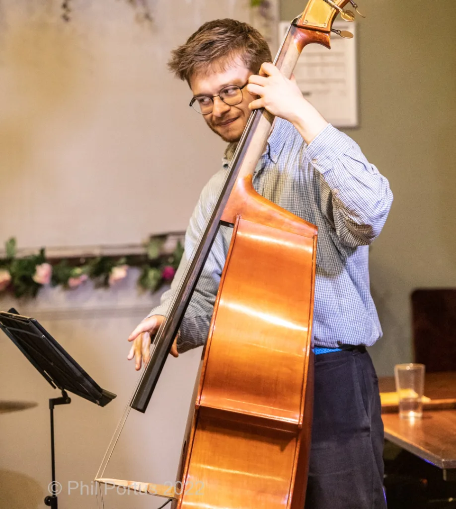 A person stands holding a double bass, wearing glasses and a blue shirt.