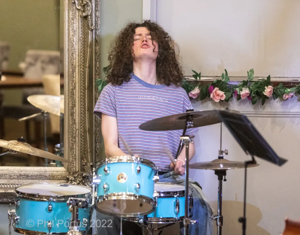 A person with curly hair plays a light blue drum kit in front of an ornate mirror and floral garland.