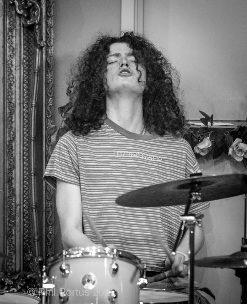A person with curly hair plays a drum set while looking upwards in black and white.