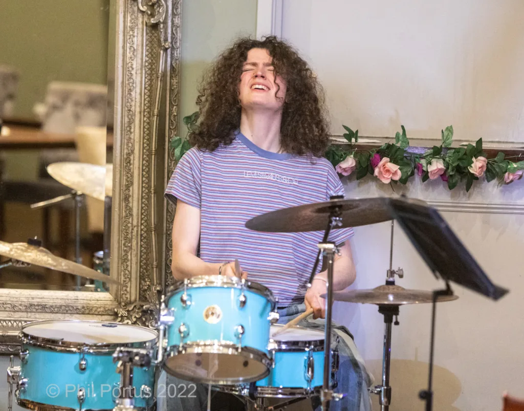 A musician with curly hair plays a blue drum kit while looking upwards.
