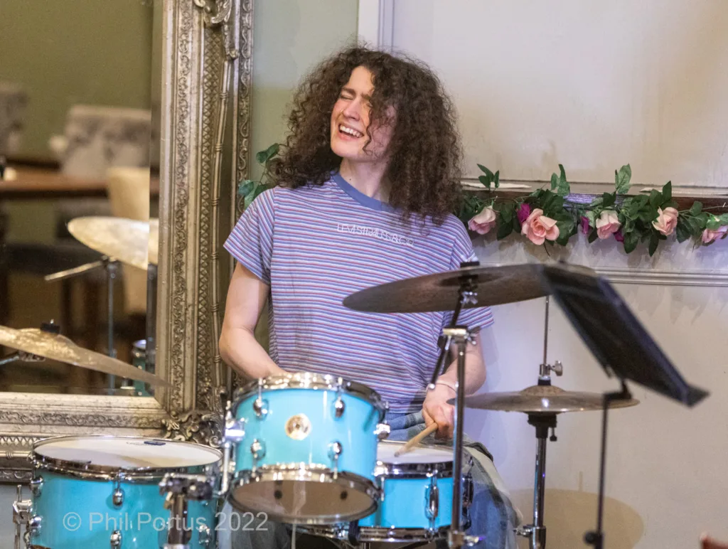 A drummer with curly hair smiles while playing a blue drum kit in front of ornate decorations.