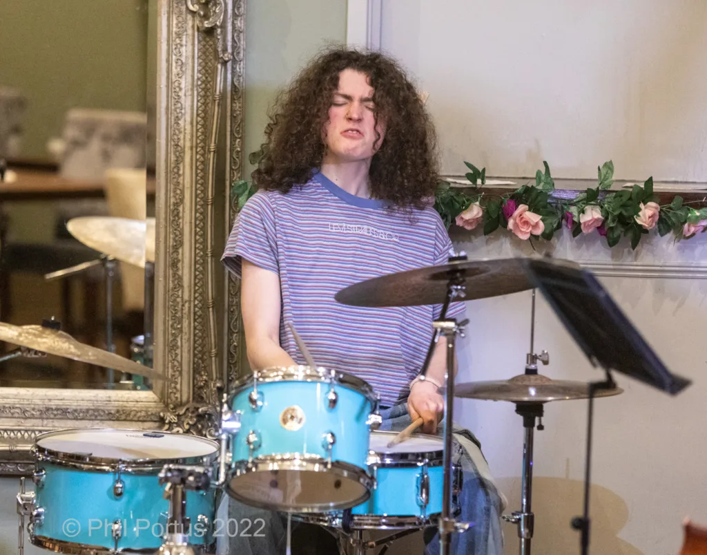 A person with curly brown hair plays a light blue drum kit in a room with ornate mirrors and floral decorations.