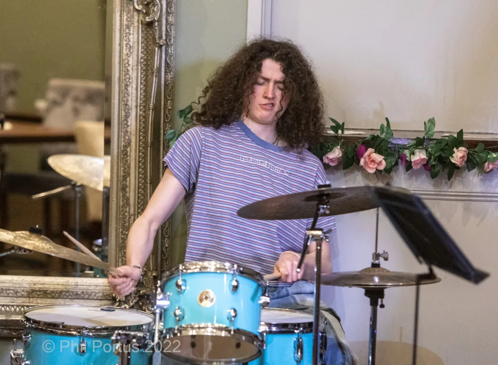A person with curly hair plays a blue drum kit while looking downwards.