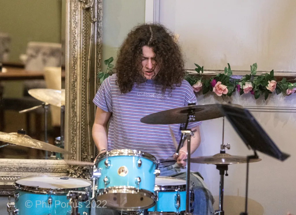 A drummer with long curly hair plays a light blue drum kit during a performance.