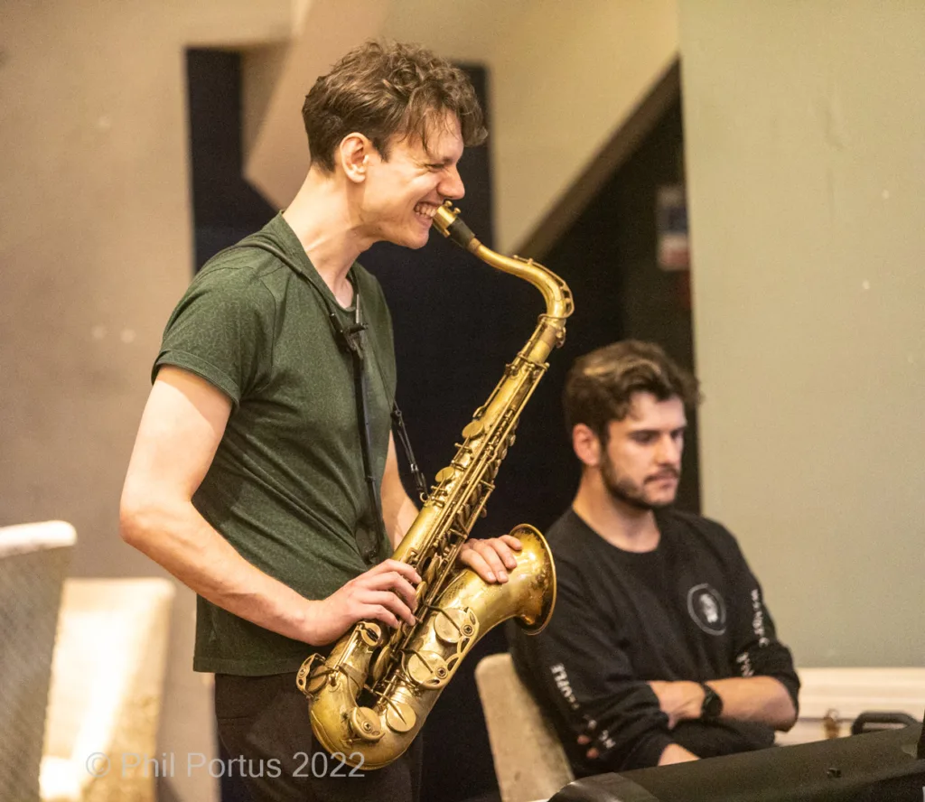 A musician plays a saxophone while smiling, with another person at a keyboard in the background.