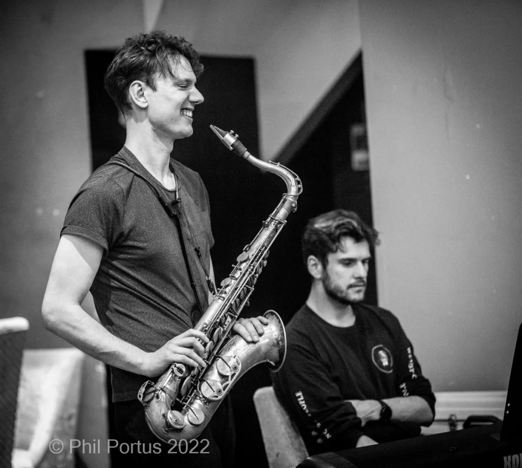 Two musicians perform on stage; one plays a saxophone while the other sits at a keyboard.