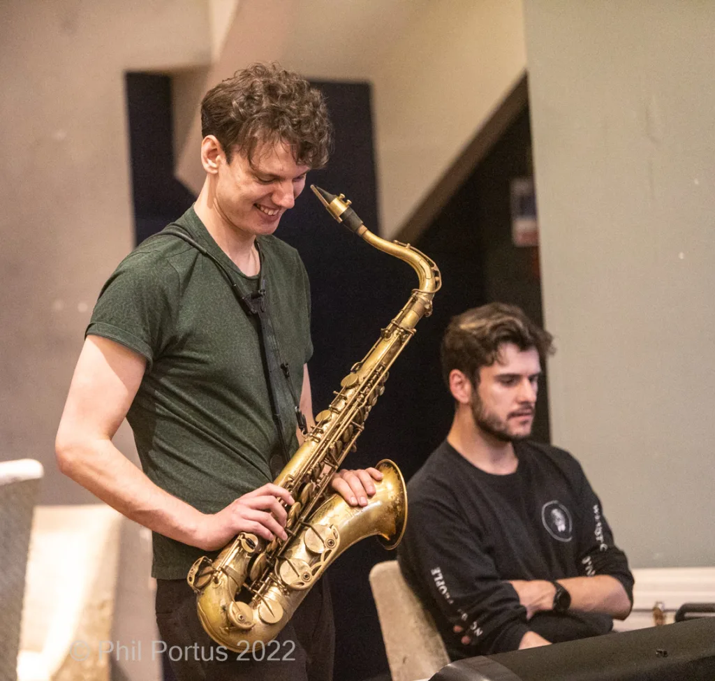 Two young men perform music; one plays a saxophone while the other sits at a keyboard.