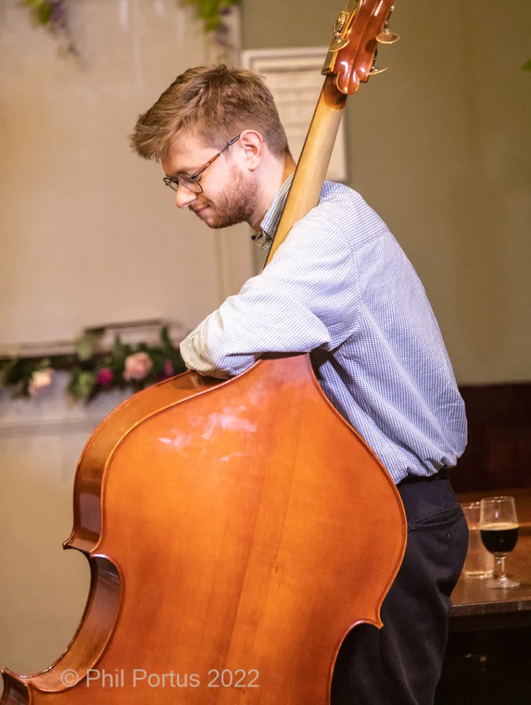 A person in a blue shirt and dark pants leans against a large double bass.