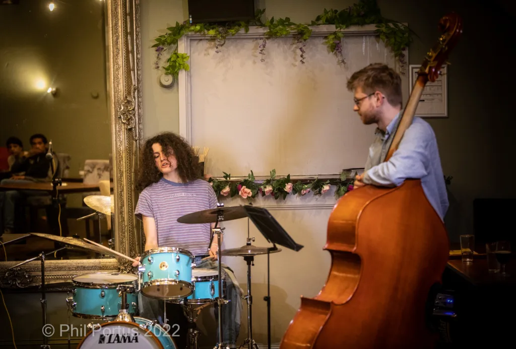 Two musicians perform on stage; one plays drums and the other a double bass.