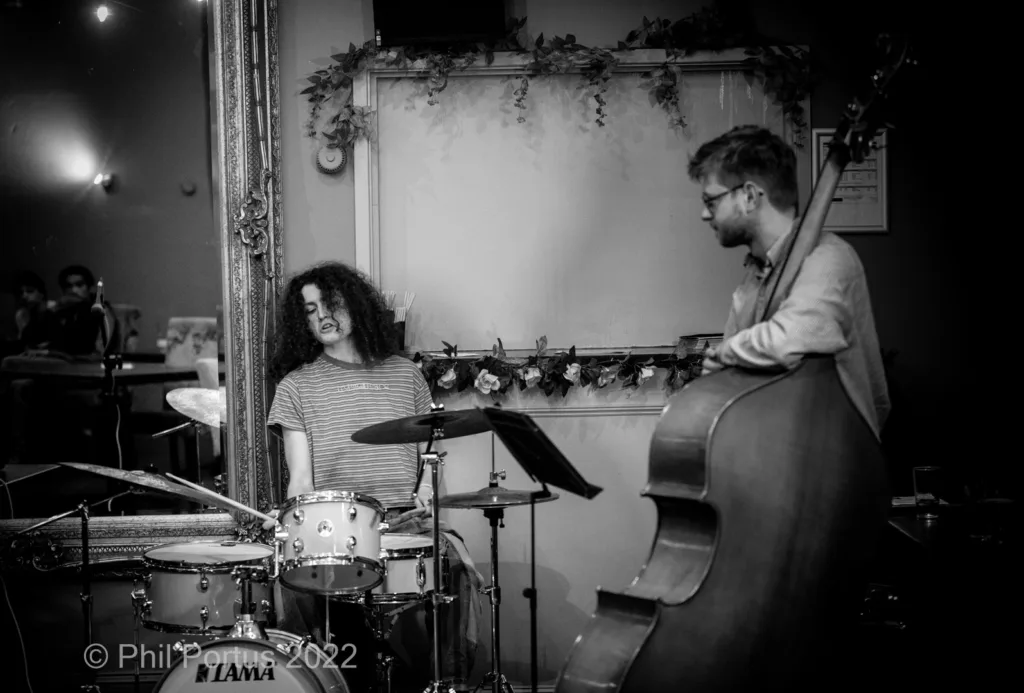 Two musicians perform on a dimly lit stage; one plays drums and the other upright bass.