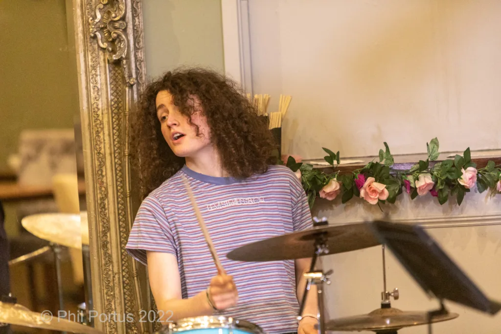 A person with curly hair plays a drum set in a room with decorative floral arrangements.