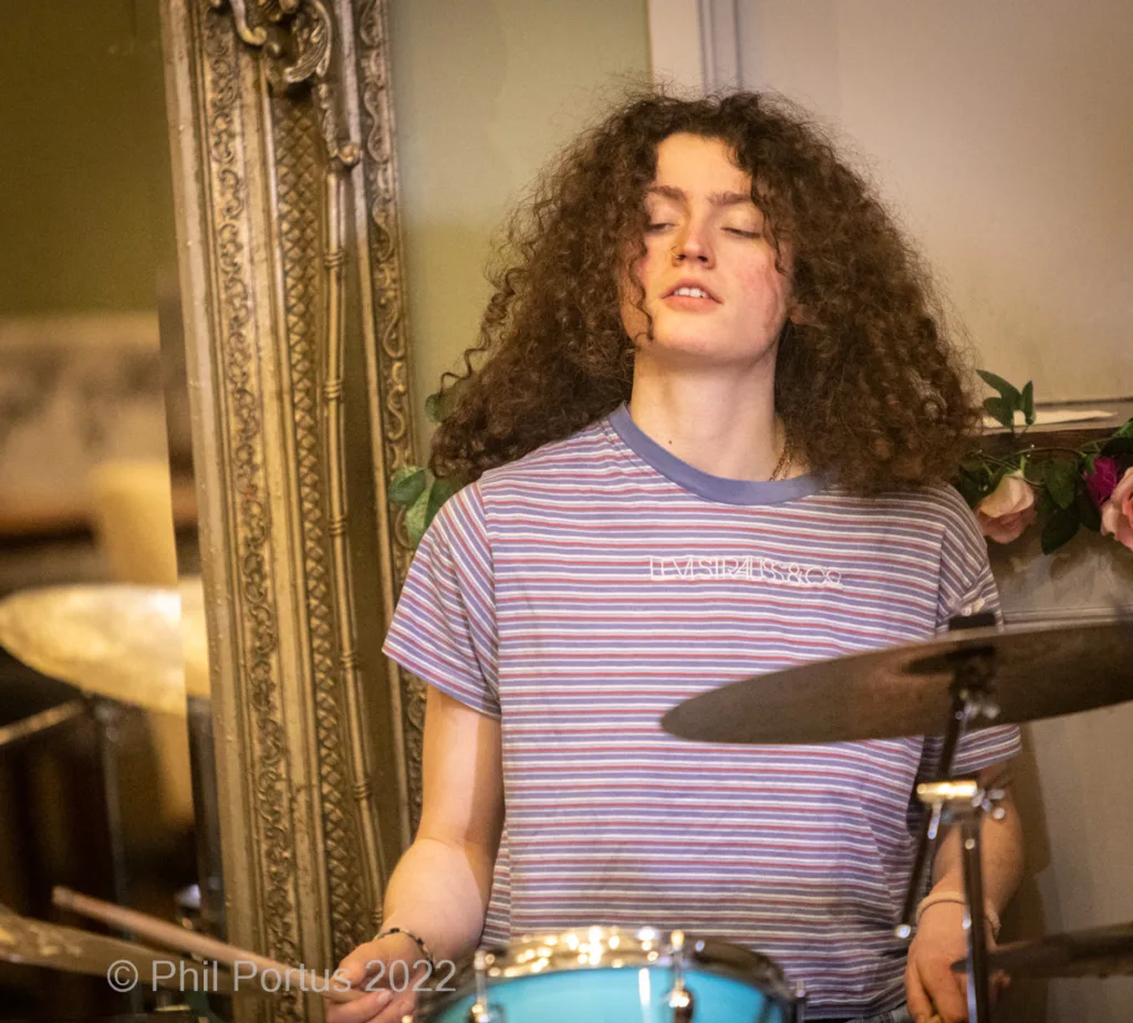 A person with curly hair plays a drum set while eyes are closed and head tilted back.