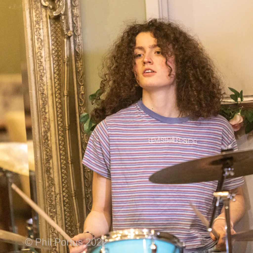 A person with curly hair plays a drum set in front of an ornate mirror.