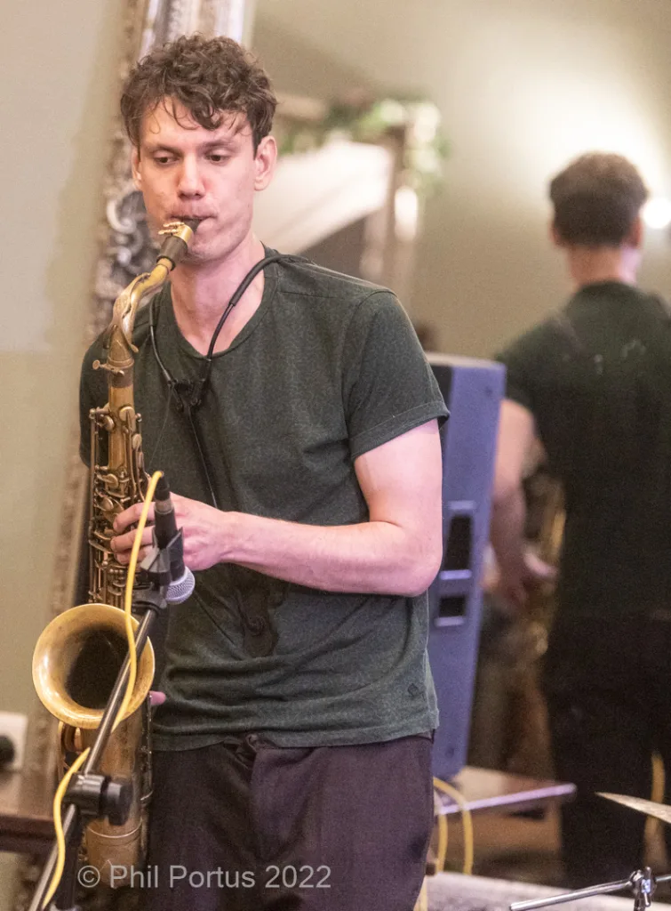 A musician plays a saxophone while standing on stage in front of a mirror and decorative plants.