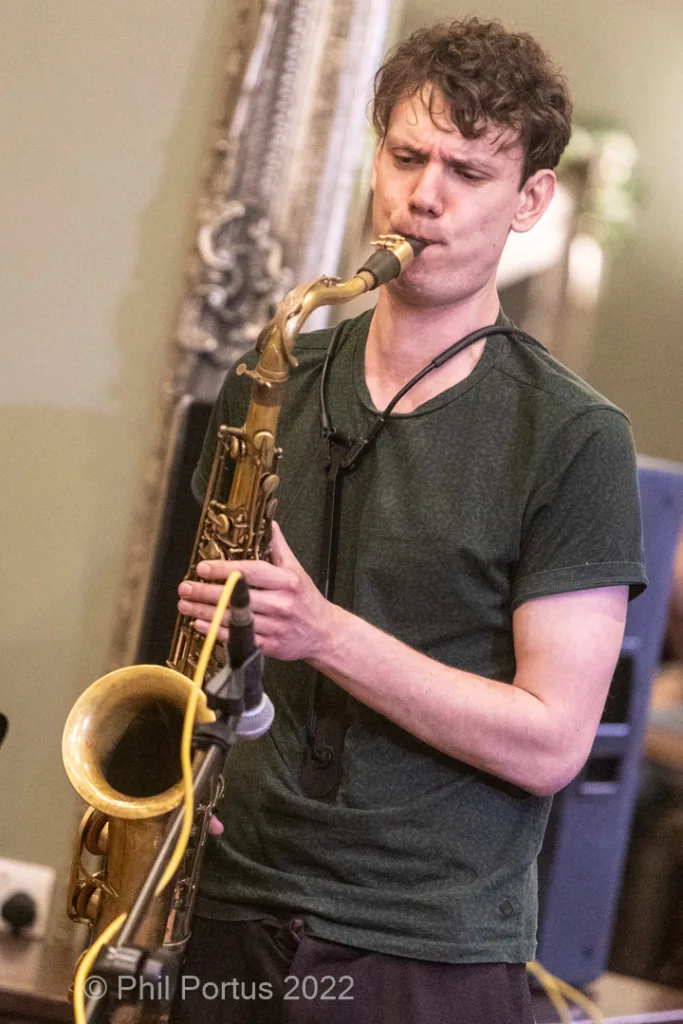 A musician plays a gold saxophone while wearing a dark green t-shirt.