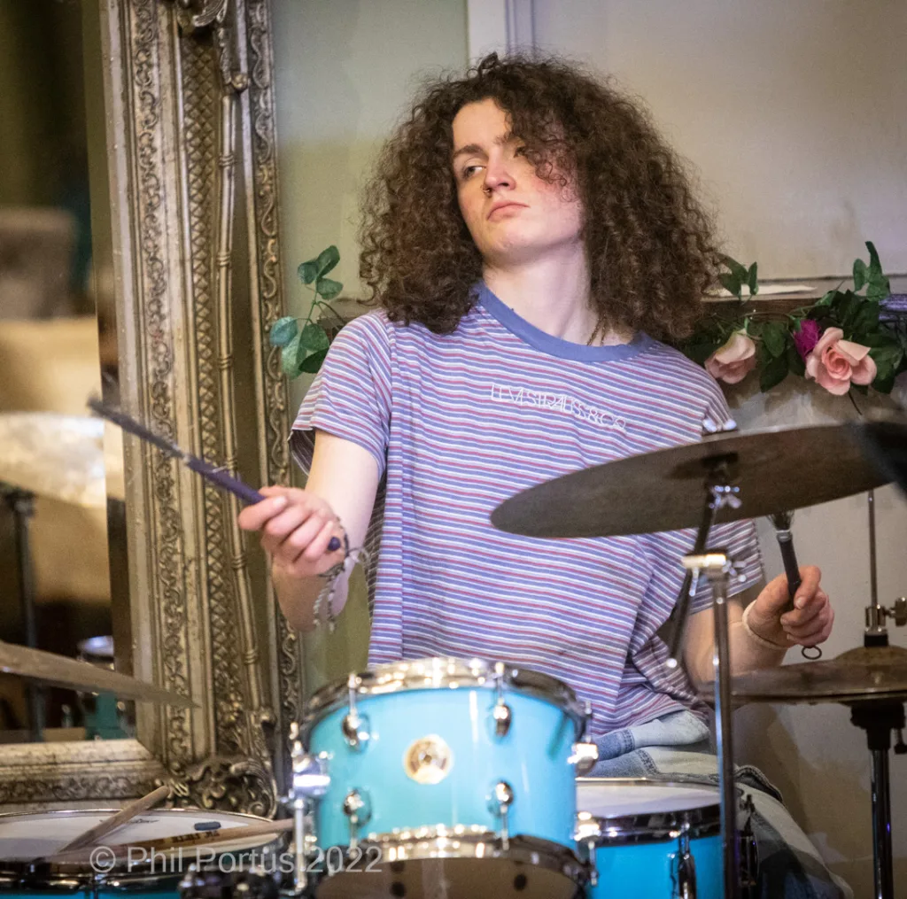A person with curly hair plays a light blue drum kit while holding drumsticks.