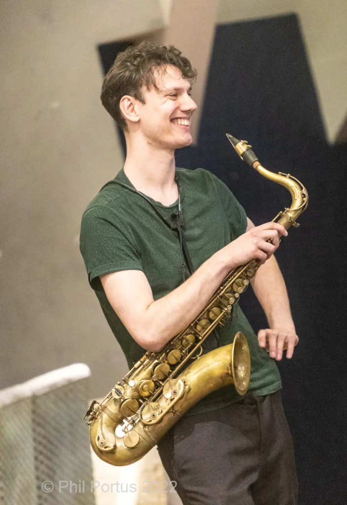 A person smiles while holding a brass saxophone on what appears to be a stage.
